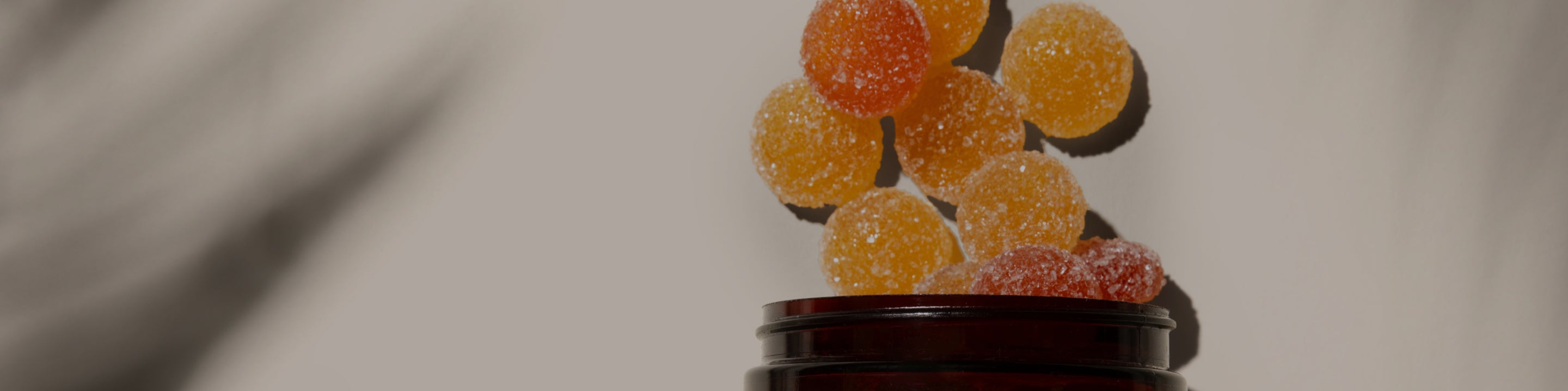 Round, Medterra gummies in various shades of orange and red are seen in mid-air above a brown jar, as if they are being poured out or are floating. The background is blurred gray, and the candies appear vibrant against it.