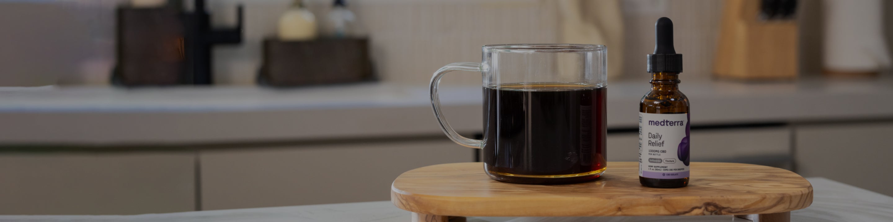 A clear glass cup filled with dark liquid is next to a small brown glass dropper bottle labeled "daily relief" on a wooden platform. The background features a modern kitchen countertop with various items out of focus.