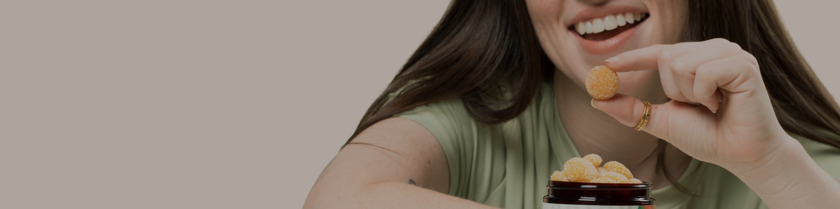 A woman with long brown hair, wearing a green shirt, is smiling and holding a Medterra gummy above a jar filled with CBD gummies. Her focus is on the gummy she is about to eat. The background is plain and light-colored.