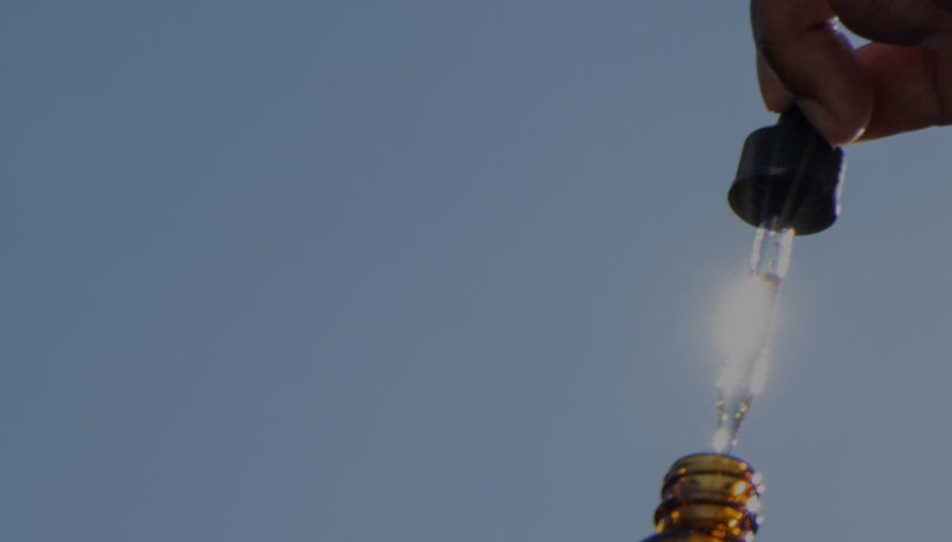 A person holds a dropper filled with liquid above a small, brown, glass bottle against a background of a clear blue sky. The close-up shot focuses on the dropper and bottle, with partial visibility of the person's hand and arm.