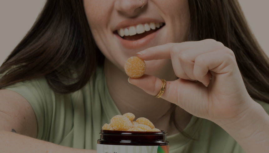 A woman with long brown hair, wearing a green shirt, is smiling and holding a Medterra gummy above a jar filled with CBD gummies. Her focus is on the gummy she is about to eat. The background is plain and light-colored.