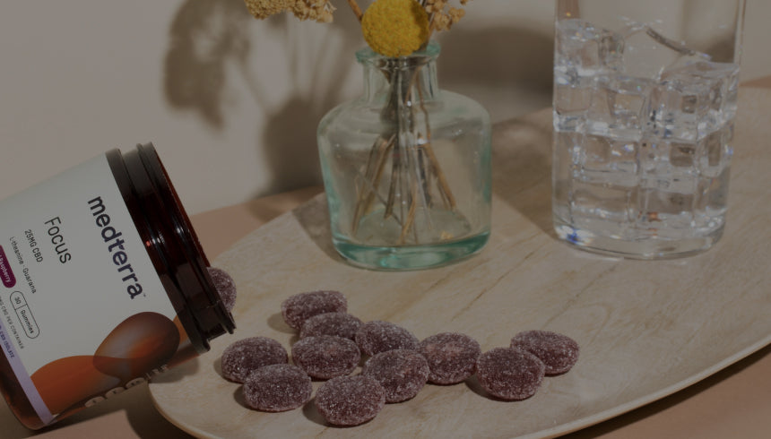 A jar of Medterra Focus gummies lies on its side, spilling gummies onto a wooden tray. Beside the tray are two glass vases with flowers and a glass of water. The background is neutral with a light beige wall and soft peach surface.