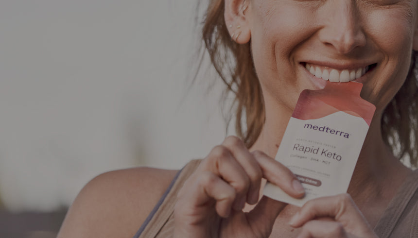 A smiling woman is holding a packaged product labeled "Medterra Rapid Keto" while standing outdoors. She has her hair tied back and is dressed in casual attire, with a blurred background of trees and a clear sky.