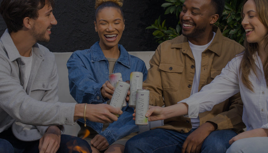 A group of four people, two men and two women, sit on a bench outdoors, smiling and clinking Medterra Delight Social Tonic cans together as they celebrate. They are casually dressed and appear to be enjoying a friendly, relaxed gathering. Green foliage is visible in the background.