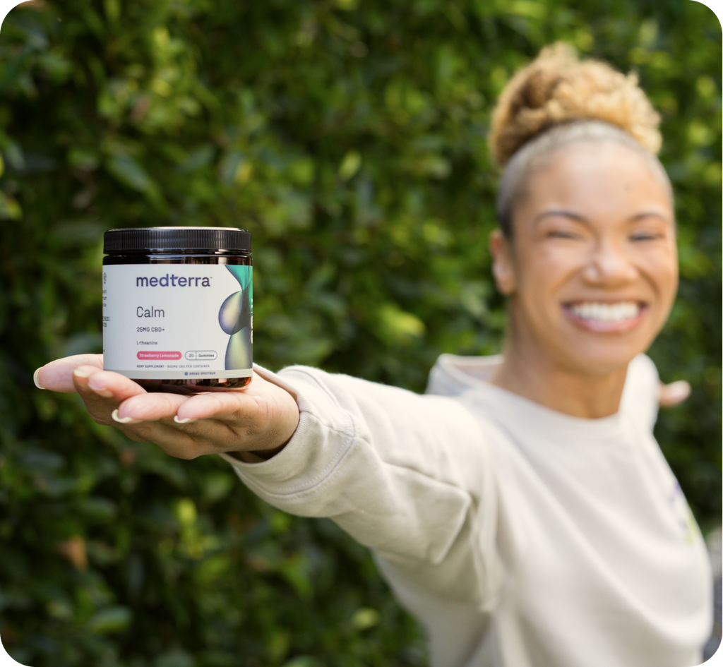 A person with a tied-back hairstyle joyfully extends their arm towards the camera, holding a jar of Medterra Calm gummies. The greenery in the background adds a natural feel to the scene, highlighting the broad-spectrum CBD's therapeutic benefits.