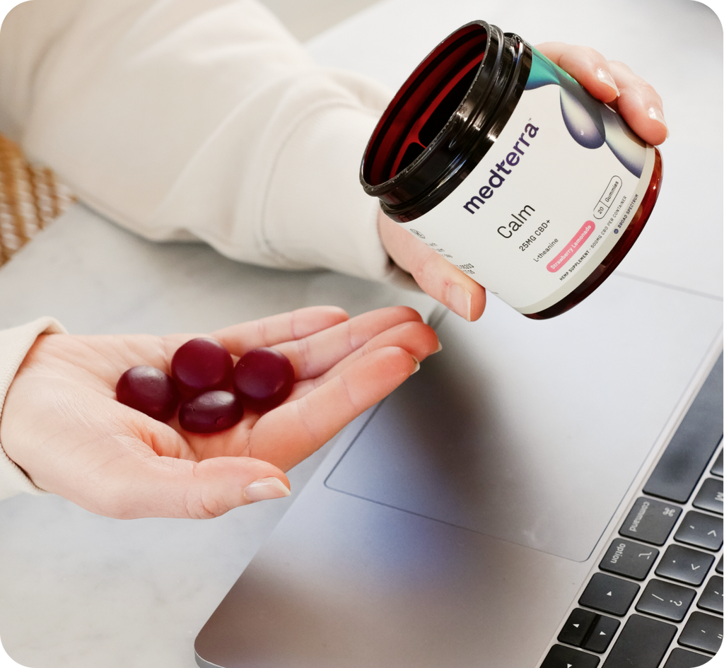 A person is sitting at a table, holding three Calm gummies in one hand and a jar labeled "calm" in the other. The jar, branded with "medterra," highlights its broad-spectrum CBD benefits. A laptop is partially visible on the table, setting a scene of modern relaxation.