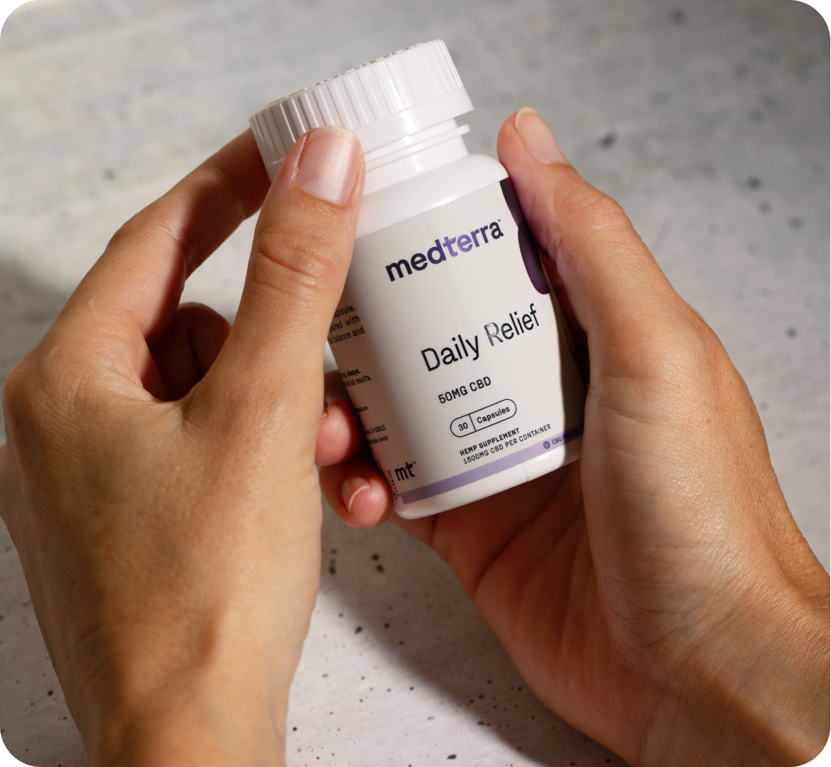 A person holding a travel-friendly white bottle labeled "medterra Daily Relief," which highlights "50MG CBD" and an icon of a capsule. This zero THC bottle is being held over a light-colored stone surface.