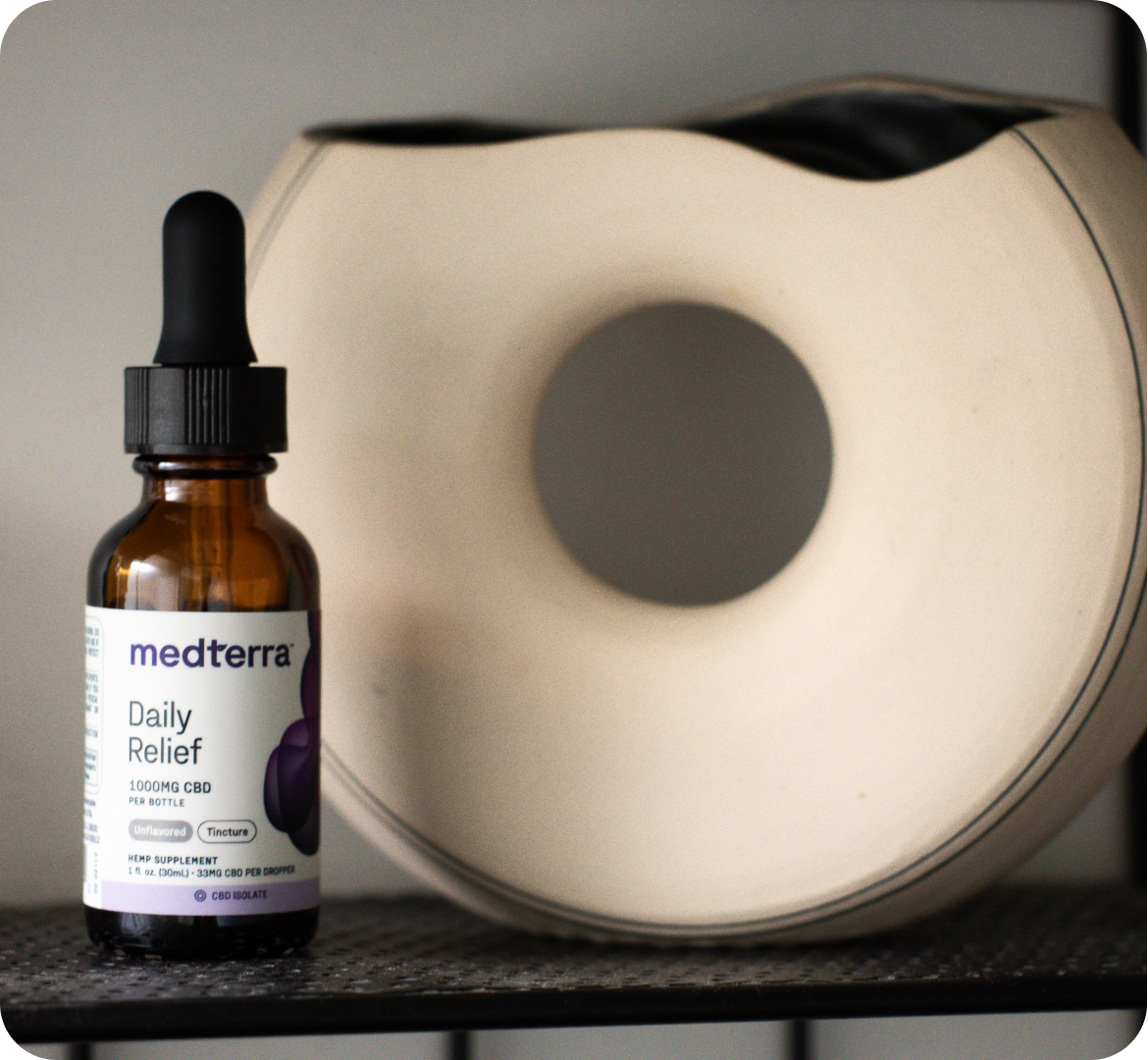 A bottle of Medterra Daily Relief CBD oil with a dropper, containing pure cannabidiol extract and MCT oil, is placed on a shelf next to a modern ceramic vase with a circular opening in the center. The bottle's label indicates it contains 1000mg of CBD. The scene has a minimalist and clean aesthetic.