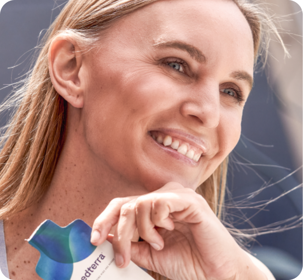 A person with long hair smiles while holding a packet with a blue and teal design, featuring the word "doTERRA." Promoting enhanced brain function, this nootropic product is aptly named Elite Memory. The background is softly blurred, directing attention to the person’s joyful expression.