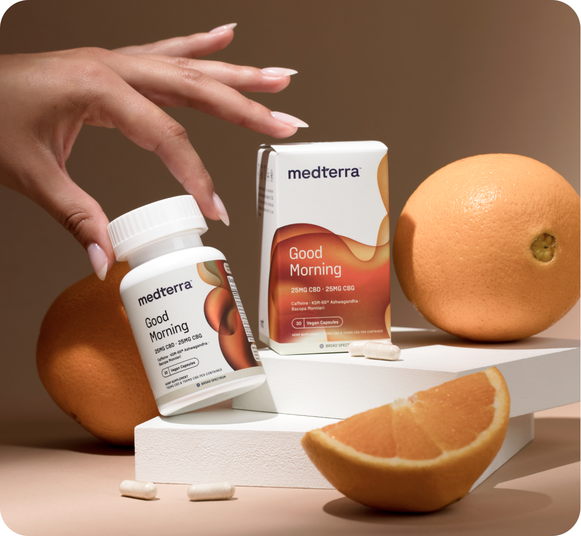 A hand reaches for a bottle of Medterra's "Good Morning" CBD supplements, placed alongside the product's packaging. A whole grapefruit, a grapefruit wedge, and three vegan capsules complement the arrangement on a two-tier white platform against a beige background.