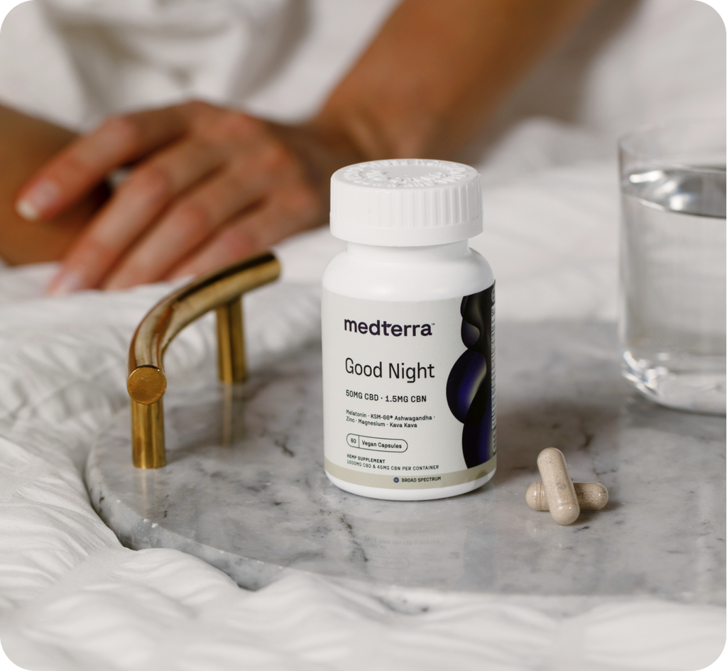 A white pill bottle labeled "medterra Good Night" rests on a marble nightstand next to a glass of water and two white Liposomal CBD capsules. In the background, a person's hand can be seen resting on a bed with white bedding, suggesting their quest for faster deeper sleep.