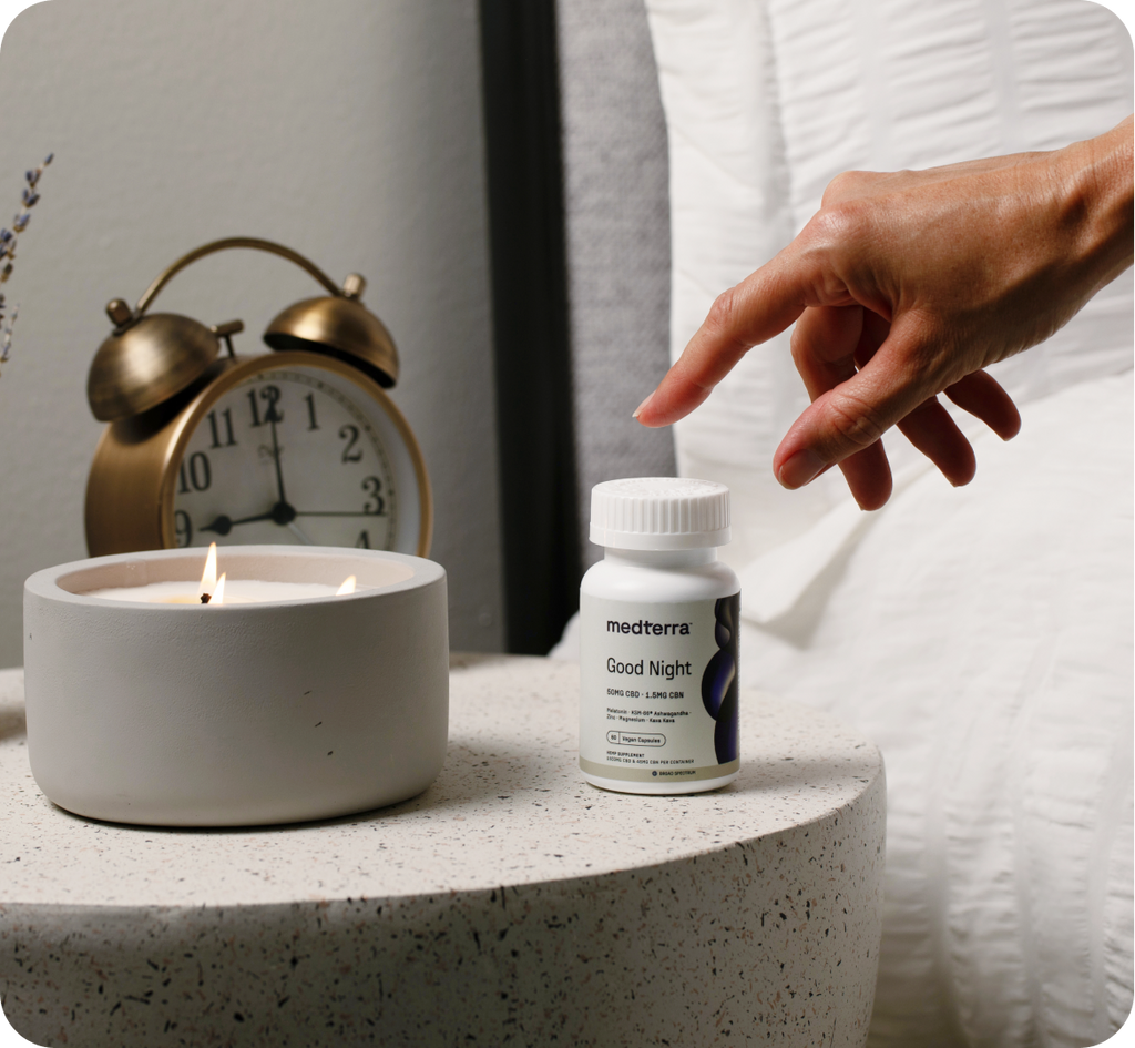 A hand reaches toward a bottle of "Good Night" capsules by medterra on a bedside table. The table also holds a lit candle and an antique-style dual-bell alarm clock displaying 10:10. Liposomal CBD capsules with melatonin promise faster sleep, while a white bed and pillow are partially visible in the background.