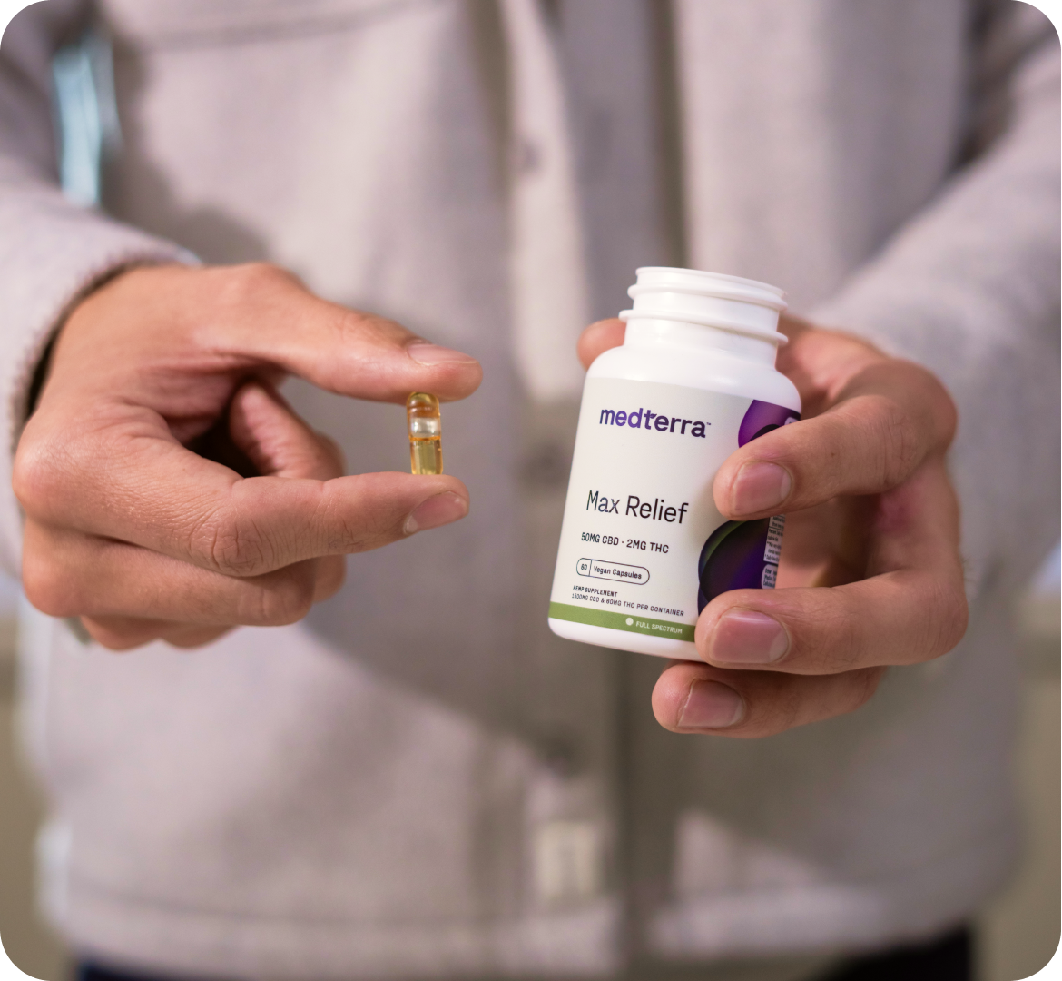 Close-up of a person wearing a light-colored jacket holding a small transparent capsule in one hand and an open white jar labeled "medterra Max Relief" in the other hand. The image suggests the use of vegan CBD capsules for relief.