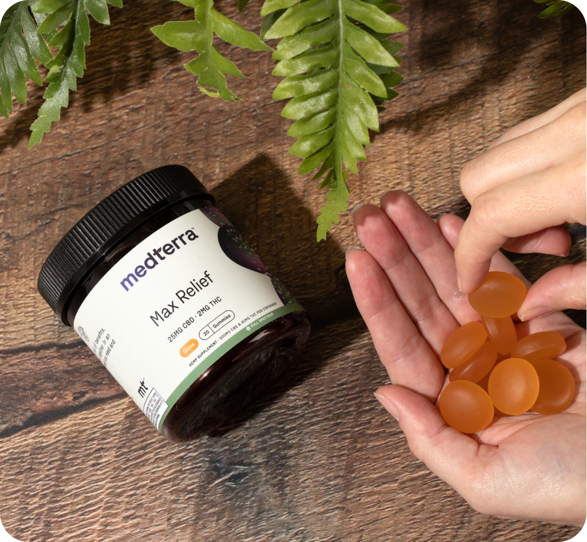A hand holding several orange, citrus-flavored gummies next to an open Medterra Max Relief CBD gummy container on a wooden surface with green leaves at the top. The container label shows "2000mg CBD + 1000mg CBG, Full Spectrum Hemp Extract.