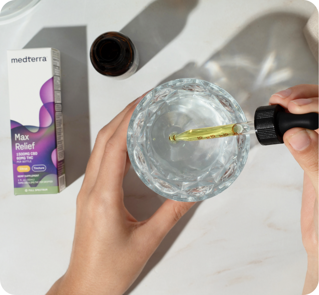 A person holds a glass of water in their left hand while using a dropper in their right hand to add liquid from a brown bottle. A Medterra Max Relief Full Spectrum CBD oil box, known for its Citrus flavors and made from organically grown hemp, is positioned on a white surface next to them.