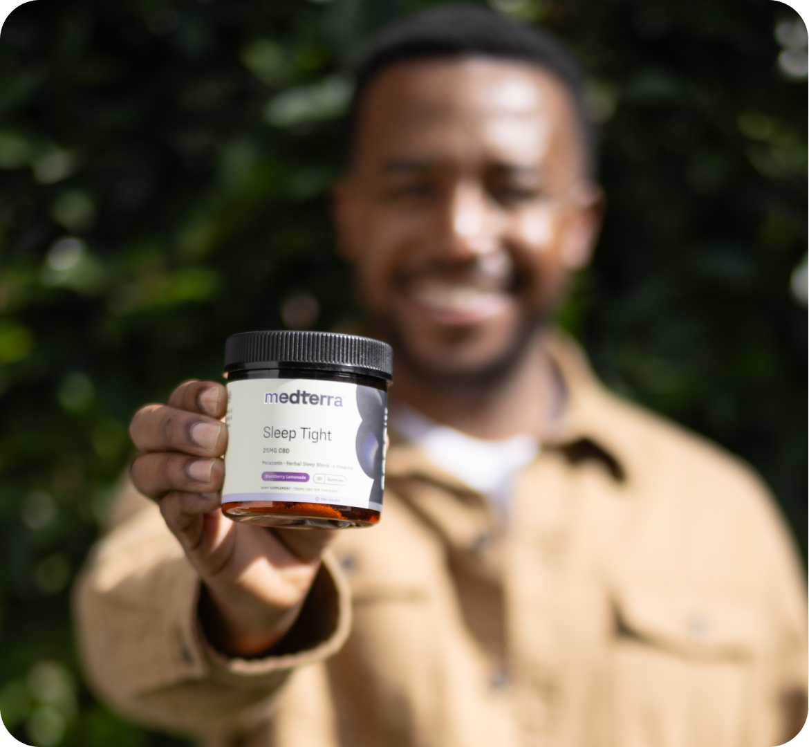 A person in a tan jacket smiles while holding a jar of Medterra Sleep Tight CBD gummies. The jar, showcasing its premium CBD and natural sleep enhancers, is the focal point, with the person blurred in the background against a leafy green backdrop.