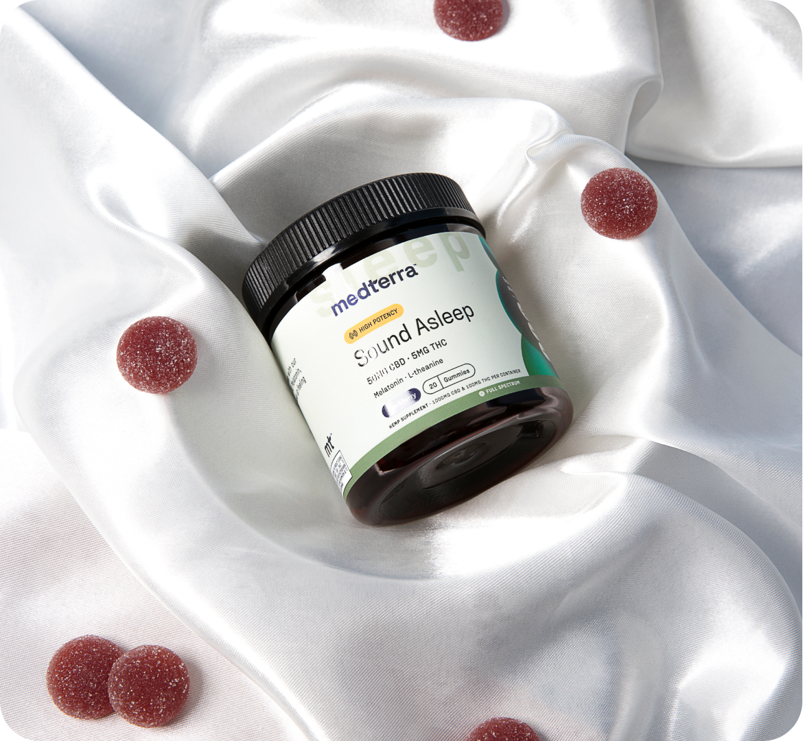 A jar of Medterra brand CBD+gummies, labeled "Stay Asleep," sits on a silky white fabric. Several red CBD sleep gummies are scattered around the jar, complementing the luxurious background. The jar and gummies are positioned to emphasize the product's relaxing attributes.