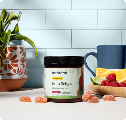 A jar of Medterra Extra Delight 10mg THC Gummies sits on a table with scattered gummies, a blue mug, a plate of orange and lime slices with raspberries, and a potted plant in front of a tiled wall.