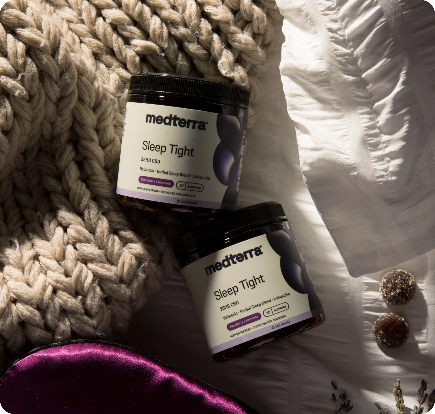 Two jars from Medterra's Multi-Spectrum CBD Sleep Bundle sit on a knitted blanket with a silk sleep mask, dried flowers, and gummies. The jars' white labels with purple accents emphasize a relaxing bedtime theme.