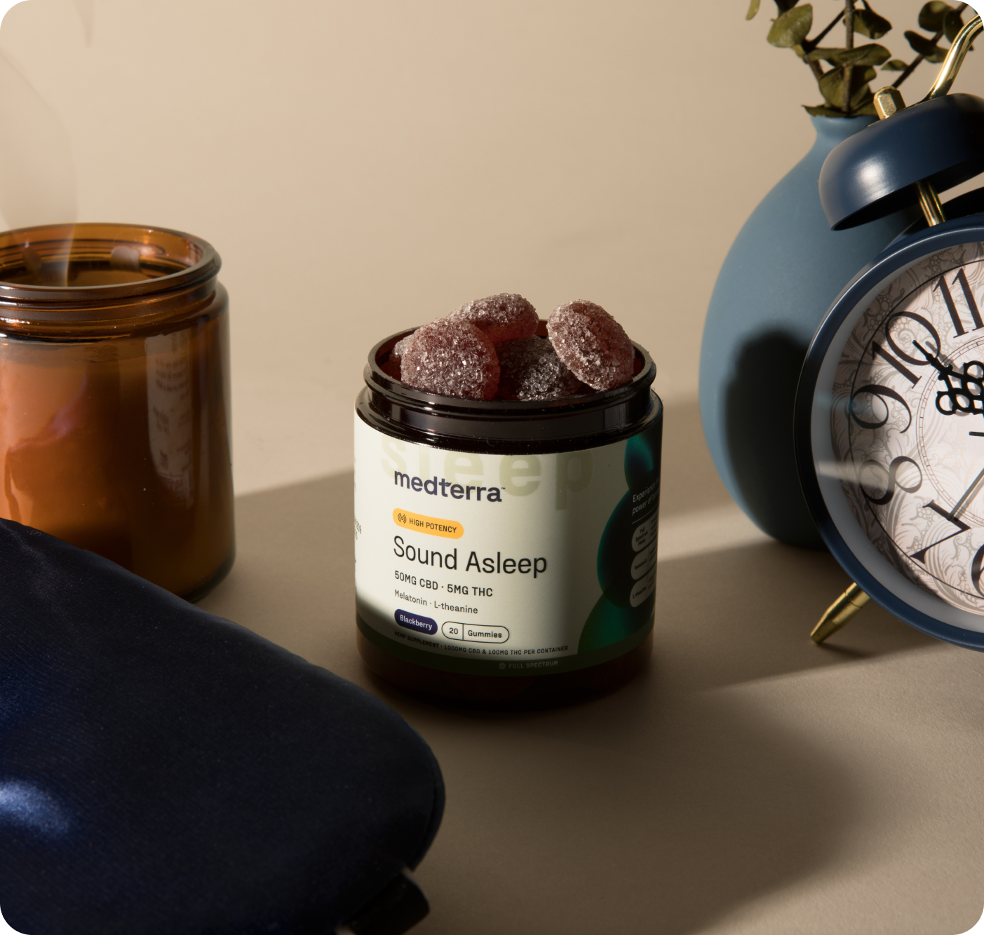 The Delight & Good Night Bundle by Medterra is open on a table, surrounded by a brown candle, a blue ceramic plant pot, a blue alarm clock, and a navy sleep mask. The setup is well-lit with soft shadows.