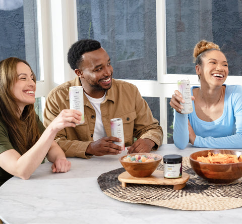 Three friends are sitting around a table, smiling and holding canned drinks. The table has snacks, including a bowl of colorful candies and chips. A small jar labeled "affordable CBD products" is also on the table. The group appears to be enjoying their time together.