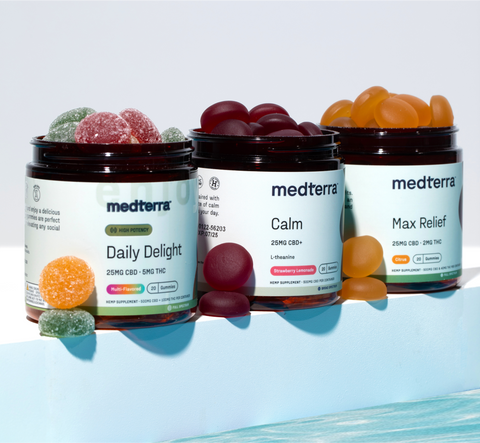 Three open jars of Medterra CBD gummies are displayed on a light blue surface. The jars, labeled "Daily Delight" with mixed fruit gummies, "Calm" with dark red gummies, and "Max Relief" with orange gummies, have some scattered around them. Perfect for any influencer or blogger highlighting their favorite CBD brand.