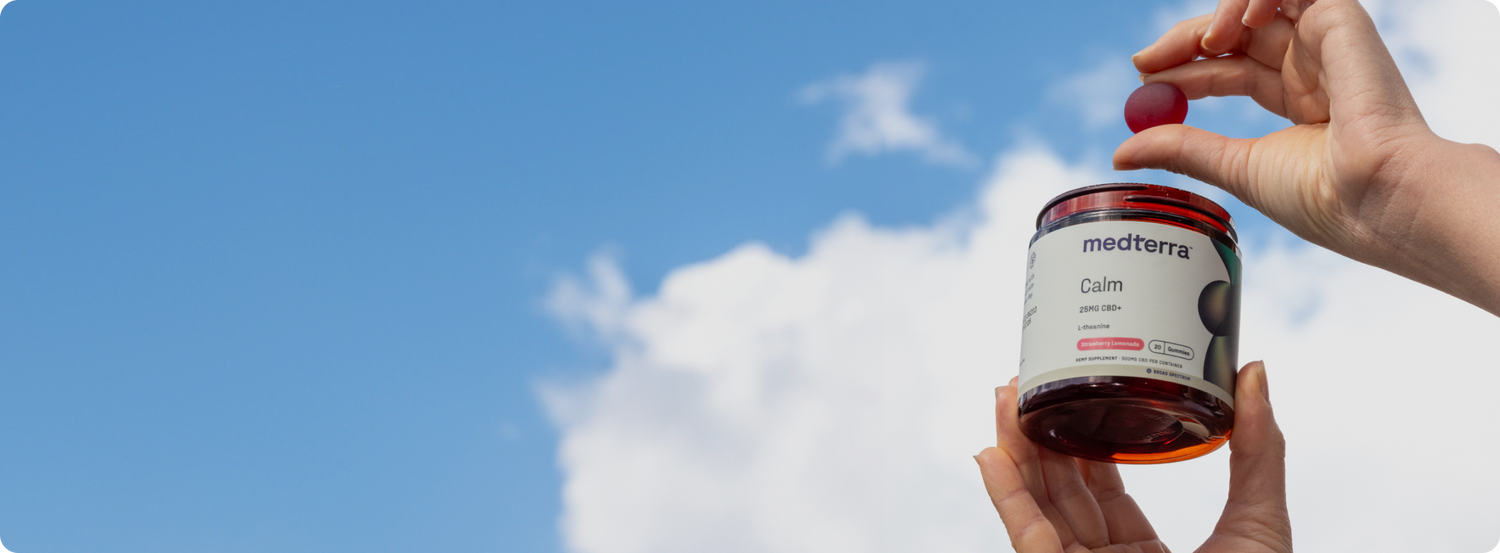 A person is holding an open jar of Medterra Calm CBD gummies with one hand, while holding a single gummy with the other hand. The background shows a clear blue sky with a few scattered white clouds.