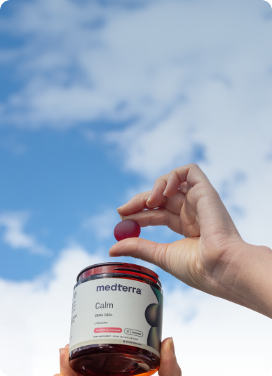 A person is holding an open jar of Medterra Calm CBD gummies with one hand, while holding a single gummy with the other hand. The background shows a clear blue sky with a few scattered white clouds.